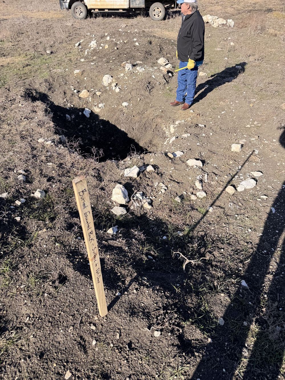 Excavated test pit with rock piles and a stake marking buried excavated rock