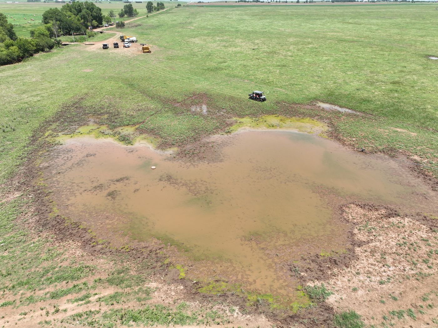 Aerial view of a large muddy pond of standing water on a pipeline right-of-way in a pasture, with inspection crew nearby